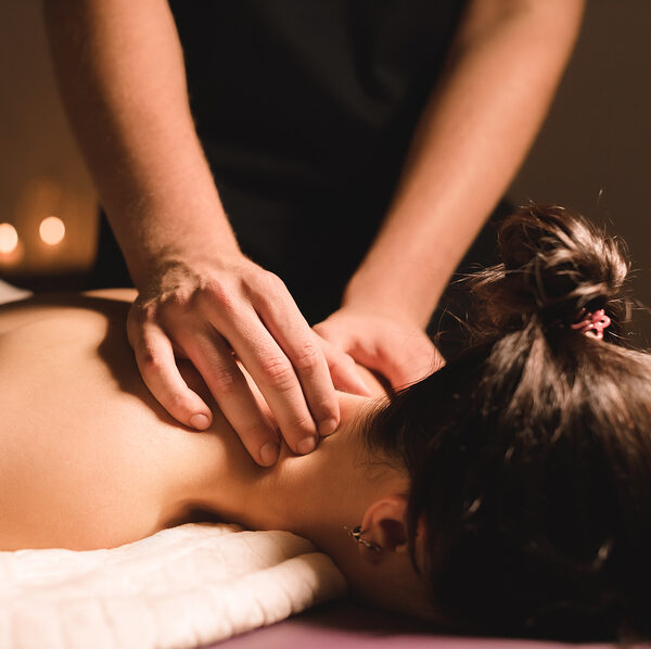 Men's hands make a therapeutic neck massage for a girl lying on a massage couch in a massage spa with dark lighting. Close-up. Dark Key.